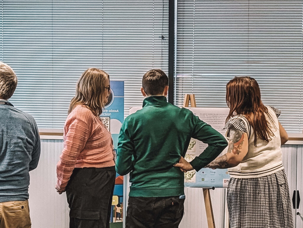 Three members of BWB staff discussing the mock consultation project around a foamex board with information on