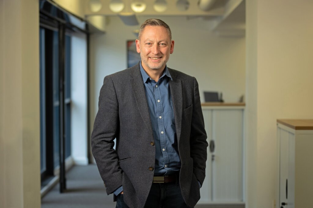 A headshot image of Simon Stanley, stood in the Leeds Office, in a walkway in between desks and window to the left